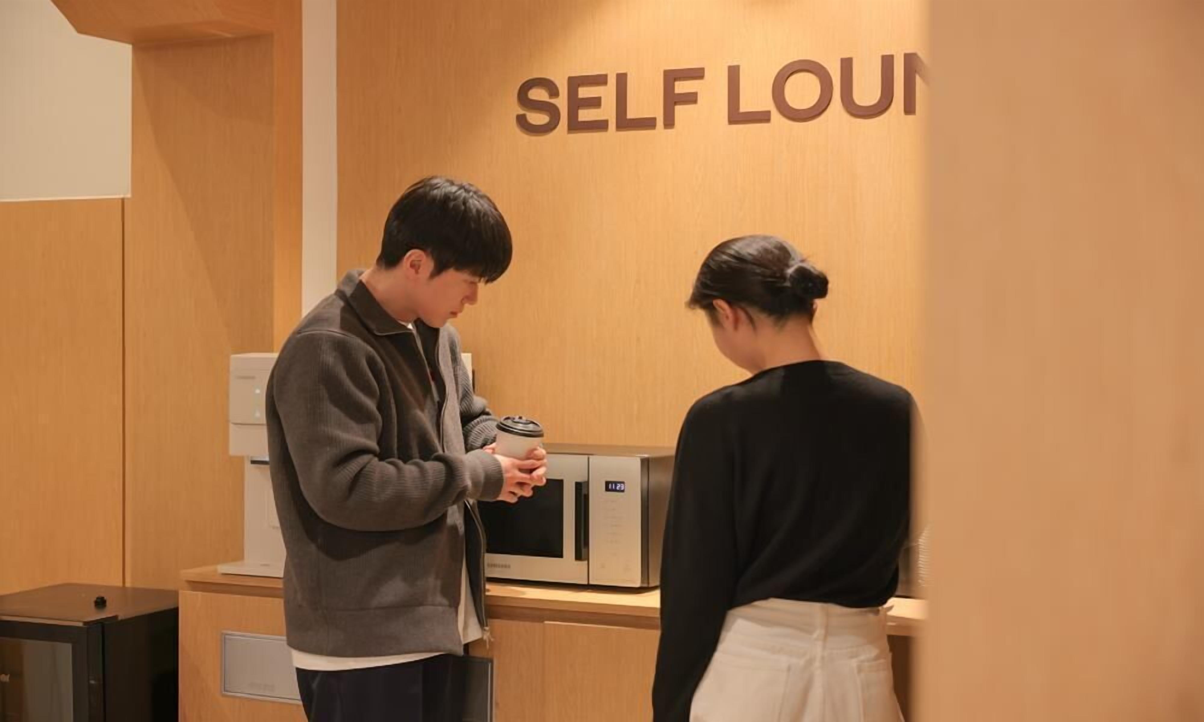 A man and a woman in a self-service lounge area with a microwave and a coffee machine.