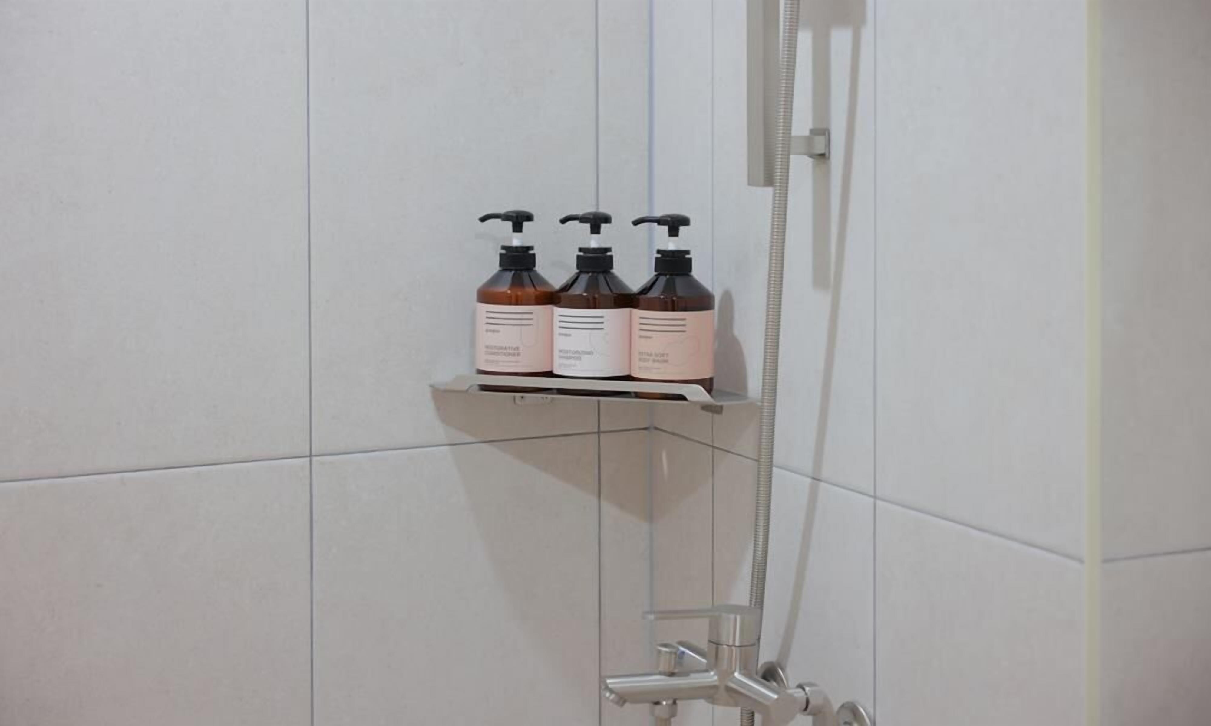 A bathroom with a shower, a glass shelf holding three bottles, and a white tiled wall.