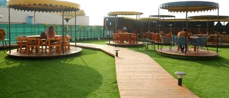 A rooftop terrace with wooden tables and chairs under canopies, surrounded by green artificial grass.