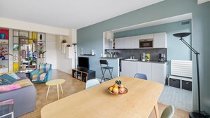 A modern living room with a sofa, a dining table with fruit, a TV, and a kitchen area with cabinets and a sink. - Appartement Akena - Welkeys (Boulogne-Billancourt)