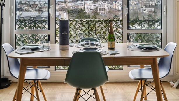 A dining table set for two with a view of a cityscape through large windows. - Appartement Akena - Welkeys (Boulogne-Billancourt)
