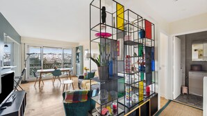 A modern living space with a large window, a dining area, and a black shelving unit with various items. - Appartement Akena - Welkeys (Boulogne-Billancourt)