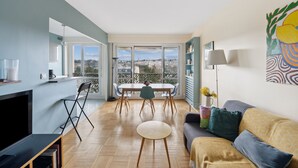 A modern living room with a dining area, large windows, and a city view. - Appartement Akena - Welkeys (Boulogne-Billancourt)