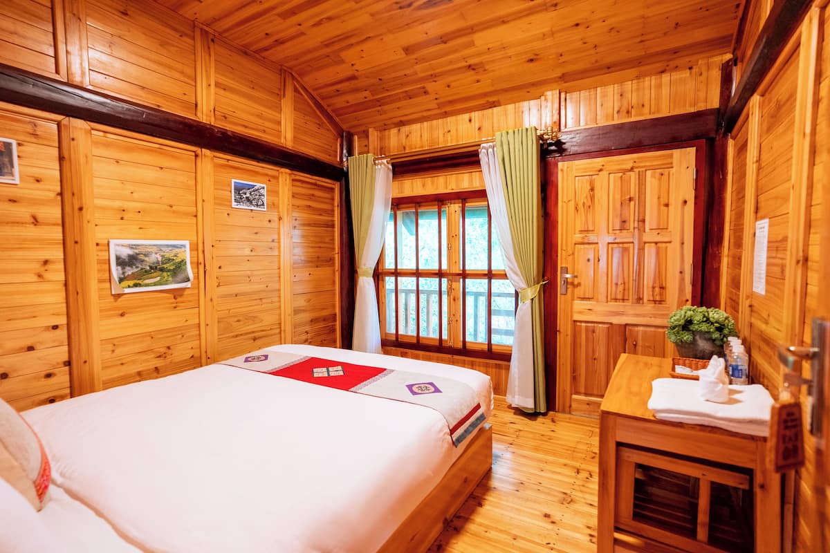 Ein Zimmer in einer Holzhütte mit einem Bett, einem kleinen Tisch mit einer Pflanze und einem Fenster mit Vorhängen.