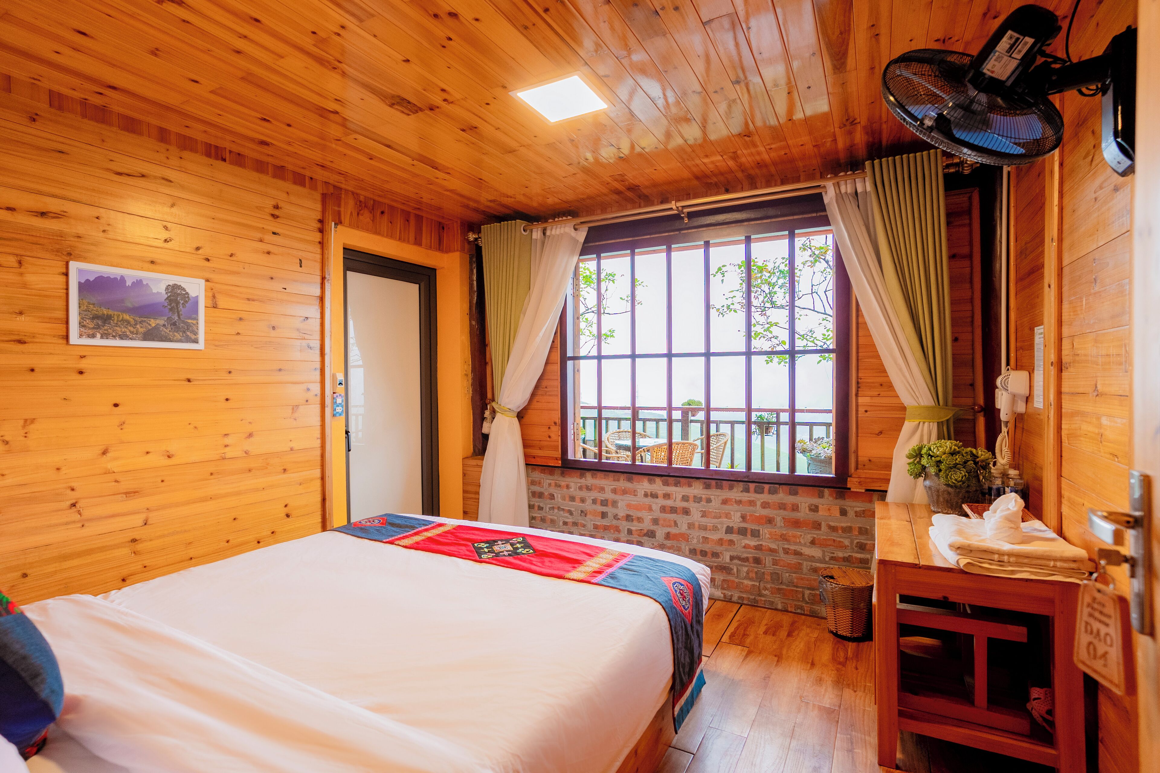Una habitación con paneles de madera, una cama, un ventanal con cortinas, un ventilador y una mesita con una planta.