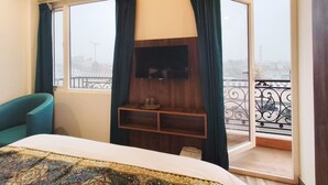 Habitación de hotel con cama, televisor montado en la pared, balcón con barandilla y vistas a la ciudad.