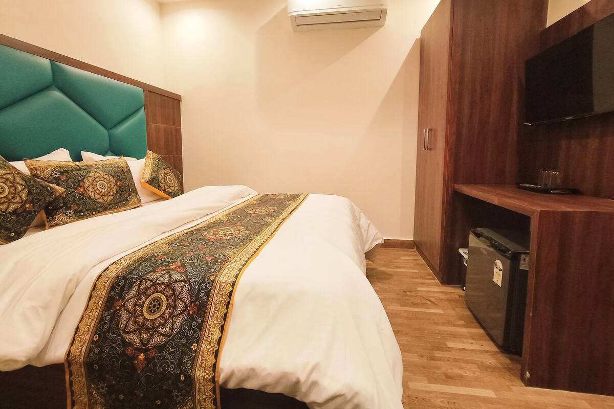 A neatly made bed with a patterned bedspread, a teal headboard, and a wooden wardrobe with a flat-screen TV.