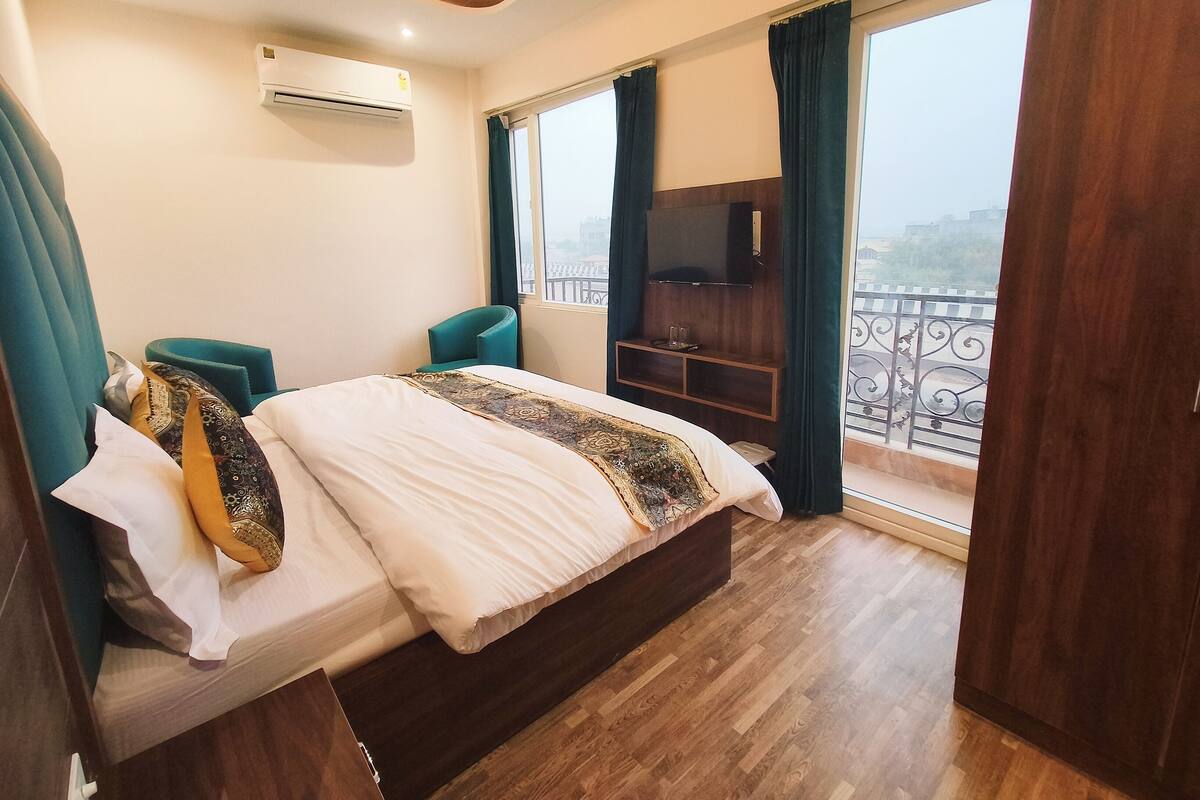 A hotel room with a large bed, a TV, a balcony with a railing, and an air conditioning unit on the wall.
