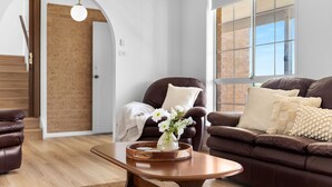 Un salon moderne avec un canapé en cuir marron, une table basse en bois ornée d’un vase de fleurs blanches et un mur en briques.