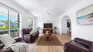 Un salon spacieux avec un canapé en cuir, une table basse et un téléviseur. De grandes fenêtres offrent une vue sur l’extérieur.