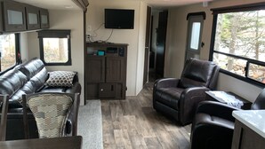 Een compact RV-interieur met een bank, een fauteuil, een aan de muur bevestigde televisie en een kleine kitchenette.