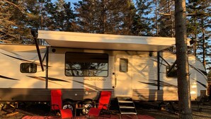 Een Forest River Wildwood RV staat geparkeerd in een bebost gebied, met een rode zeil op de grond en twee rode stoelen.