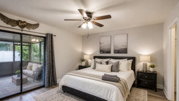 Une chambre à coucher avec un lit, un ventilateur de plafond, une table de chevet, une lampe et une vue sur l’extérieur, visible à travers une porte vitrée.