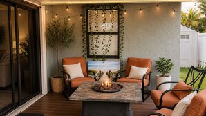 Une terrasse extérieure avec une table autour d’un foyer, deux chaises orange, une plante en pot et des guirlandes lumineuses.