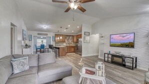 Un salon moderne avec un canapé d’angle gris, un ventilateur de plafond, un téléviseur à écran plat fixé à un mur texturé, et une cuisine en arrière-plan.