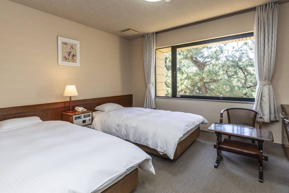 Hotelkamer met twee bedden, een bureau, een stoel, een lamp en een raam met uitzicht op bomen.