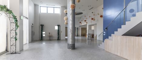 A spacious lobby with a high ceiling, marble flooring, and a central column. There are decorative elements on the ceiling and a staircase on the right.