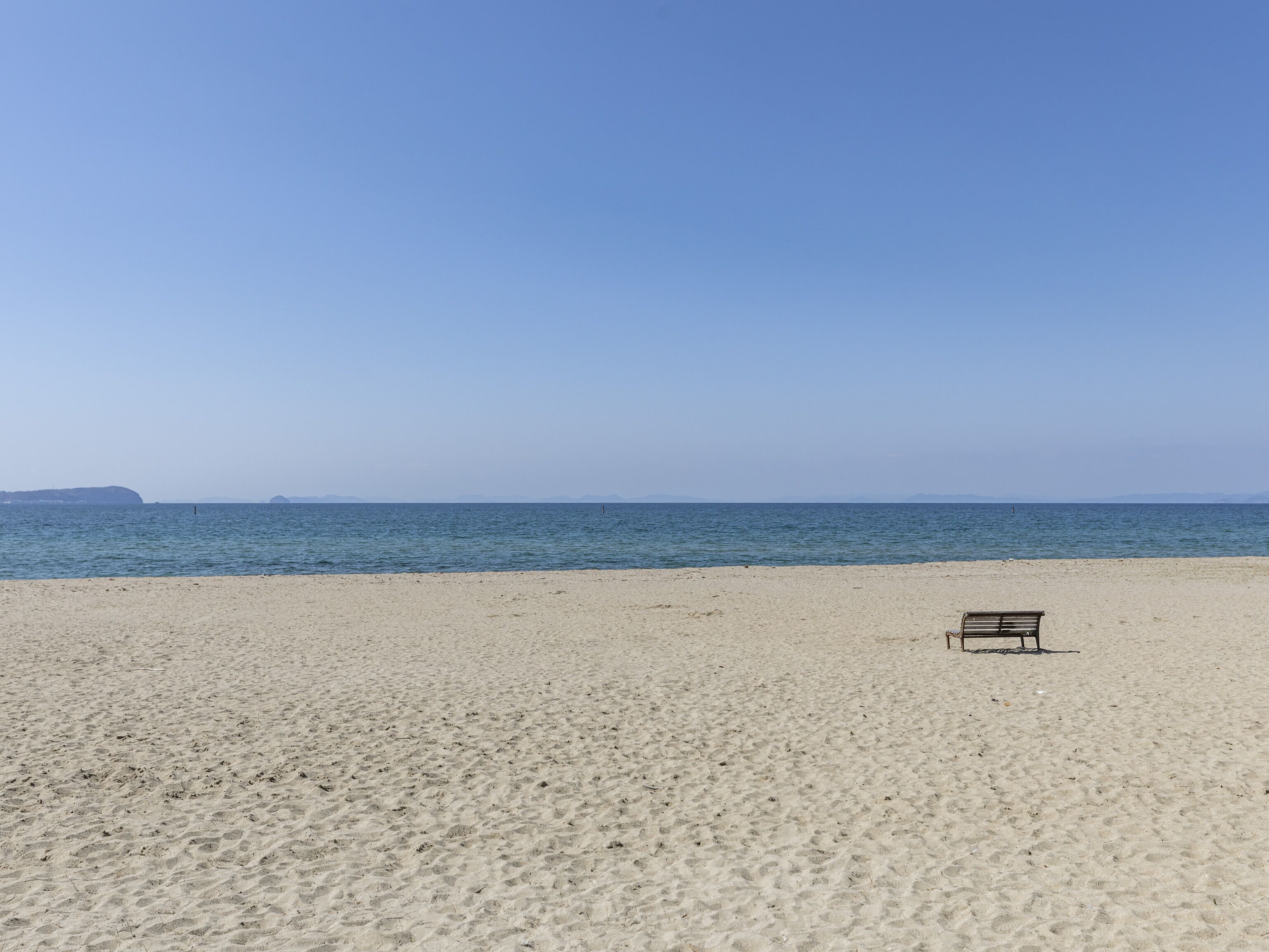 A sandy beach with a single bench and calm sea under a clear sky.
