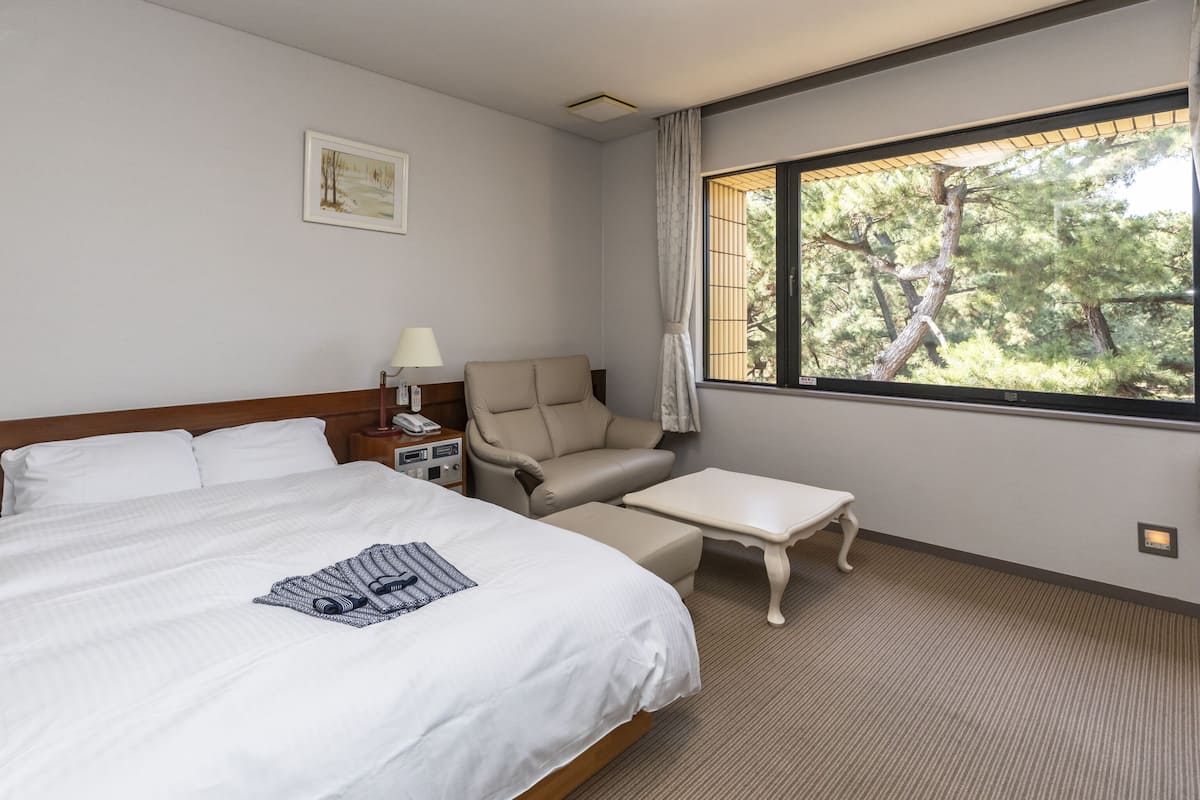 Een hotelkamer met een groot bed, een stoel, een kleine tafel en een raam met uitzicht op bomen.