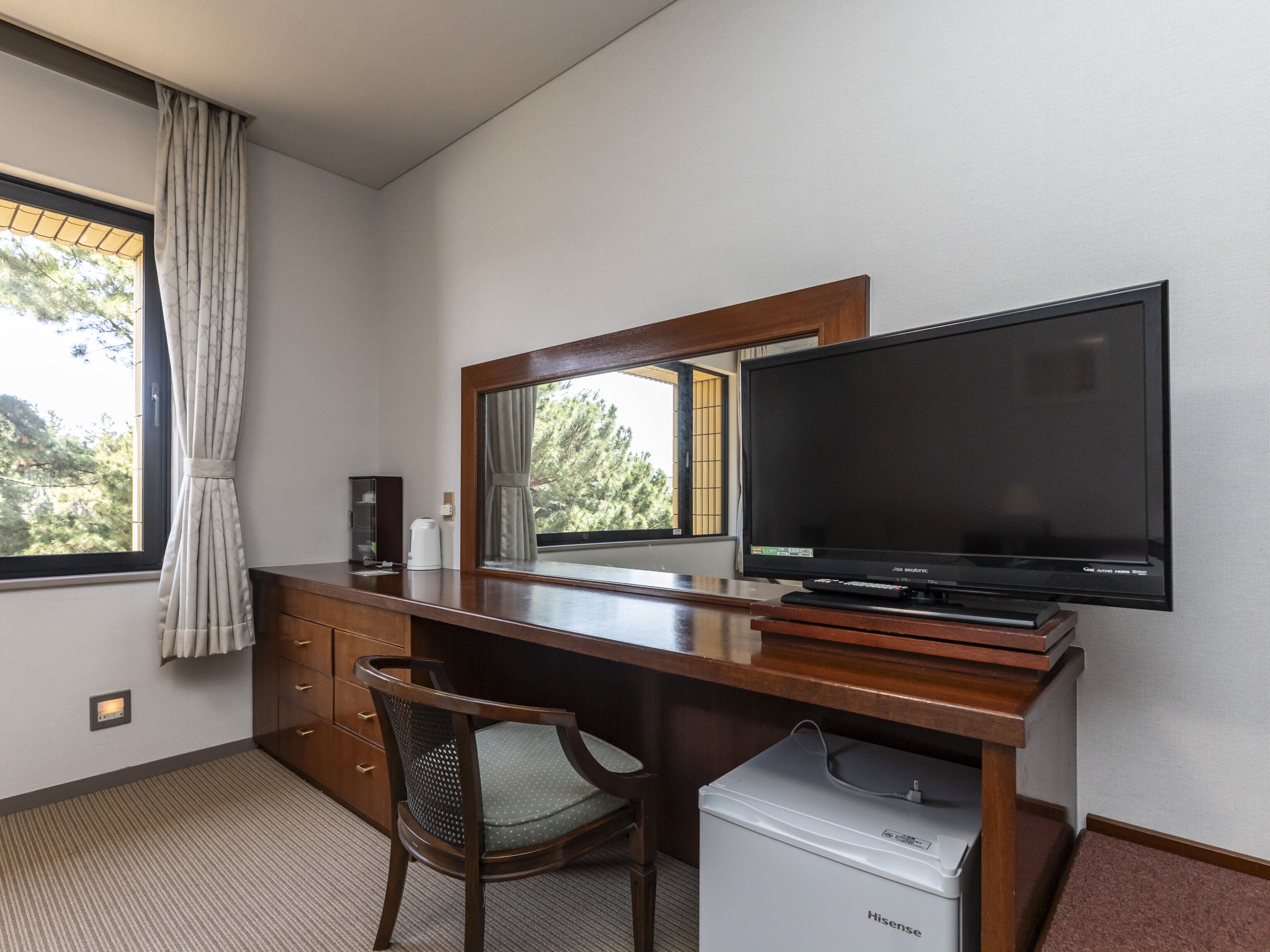 Habitación de hotel con un escritorio de madera, televisor, silla, refrigerador pequeño, ventana con cortinas y vista a árboles.