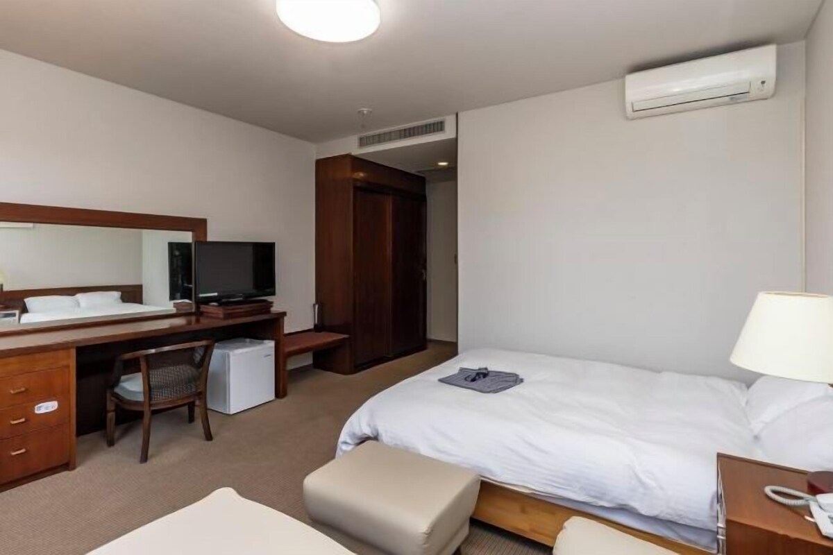 A hotel room with a bed, a desk with a chair, a television, a small refrigerator, and an air conditioning unit.
