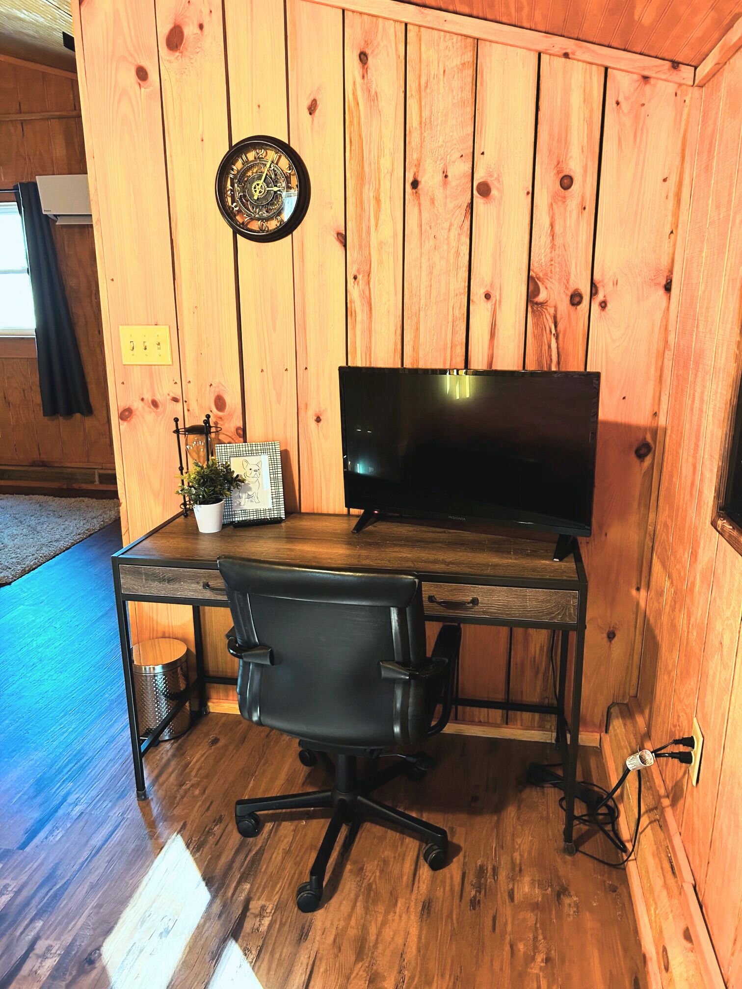 Ein Zimmer mit Holzwänden, einem Schreibtisch, einem schwarzen Bürostuhl, einem Fernseher und einem kleinen Topfpflanzen.