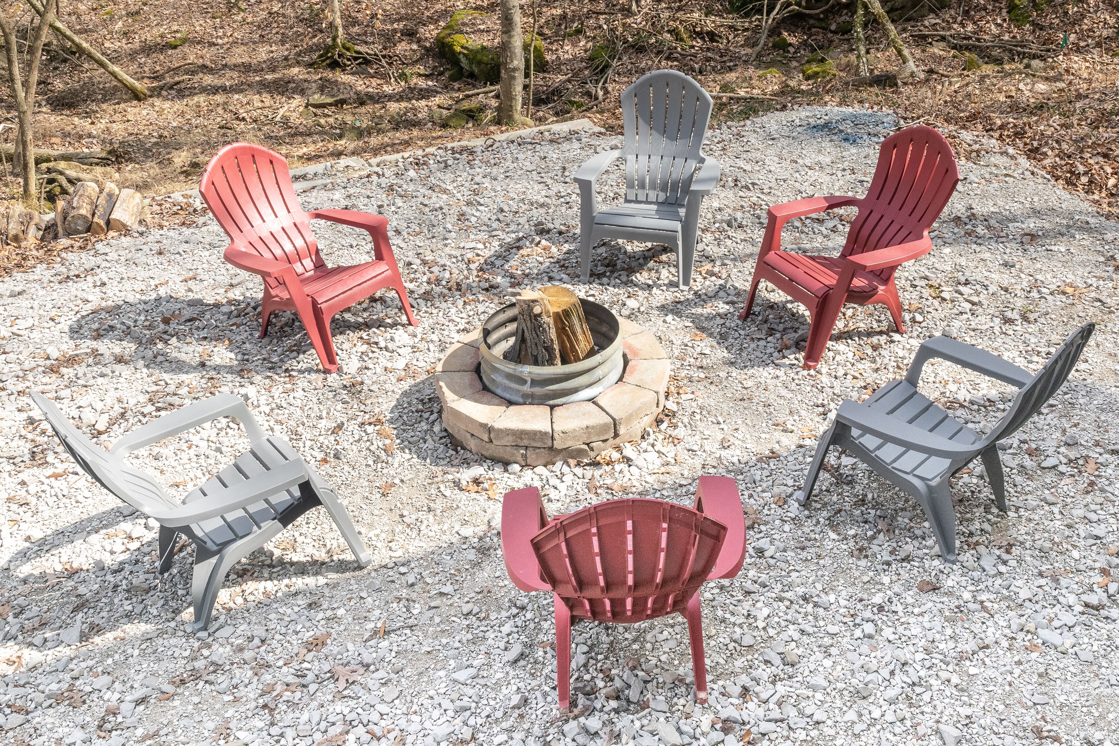 A fire pit surrounded by six Adirondack chairs in a gravel area.