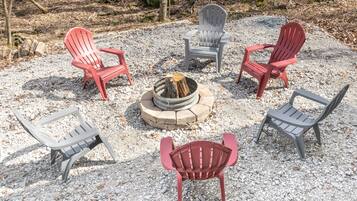 A fire pit surrounded by six Adirondack chairs in a gravel area.