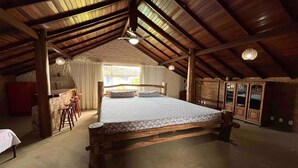Amplio dormitorio en ático con una cama grande, vigas de madera, un rincón comedor con taburetes y vistas al exterior por una ventana.