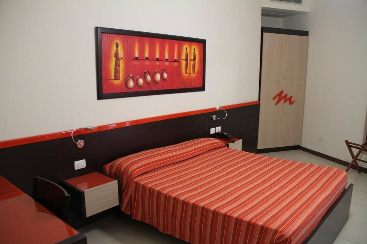 Une chambre à coucher avec un lit, des tables de chevet, une couvre-lit à rayures rouges, une œuvre d’art encadrée au mur et une armoire ornée d’un logo « M » rouge.