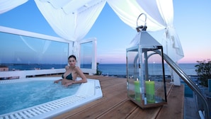 Une femme en bikini, sur une terrasse en bois avec un jacuzzi, d’où l’on a une vue sur la mer.