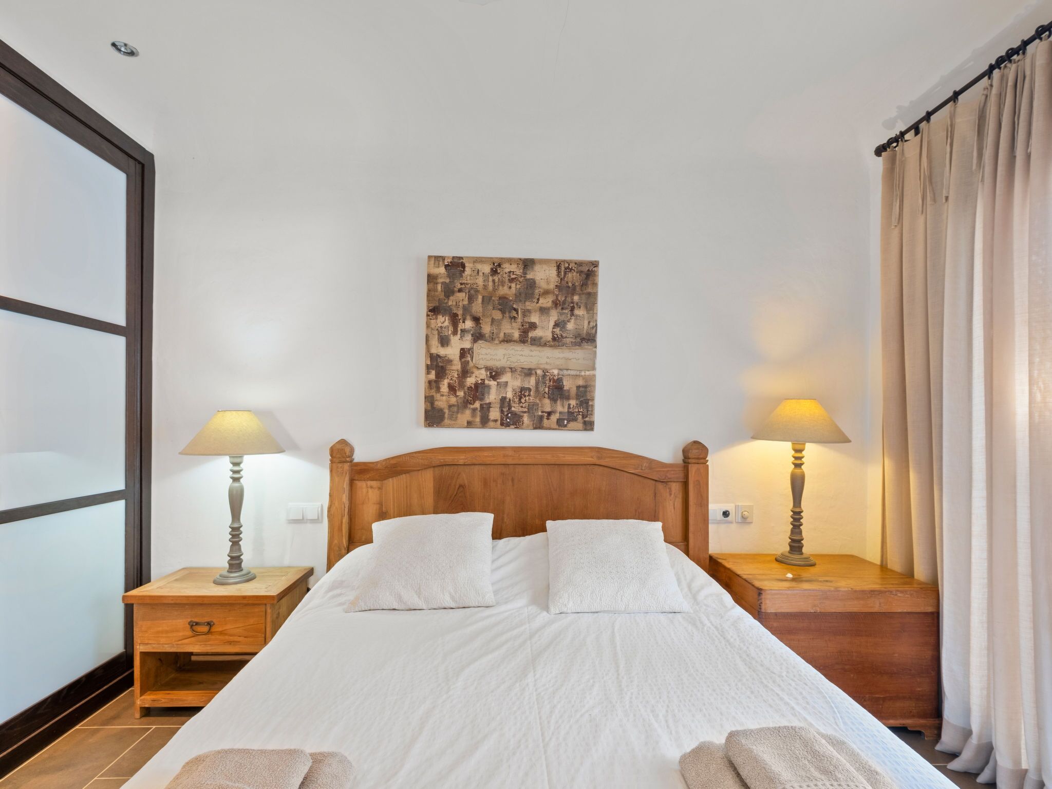 A neatly made bed with white linens, two bedside lamps, a wooden headboard, a wall-mounted artwork, and a large window with curtains.