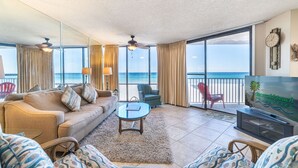 A spacious living room with a large sofa, a coffee table, and a television. The room has large windows with a view of the ocean.
