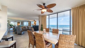 A spacious living area with a large dining table, wicker chairs, a ceiling fan, and a view of the ocean through large windows.