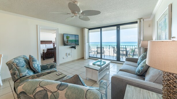 A spacious living room with a ceiling fan, a large window with a view of the beach, and a flat-screen TV mounted on the wall. - Beach Days Beachfront-Edgewater Resort in PCB 2nd Floor-3/3-Amazing! (Panama City Beach)