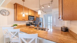 A modern kitchen with wooden cabinets, a granite countertop, white chairs, and a clock on the wall. - Beach Days Beachfront-Edgewater Resort in PCB 2nd Floor-3/3-Amazing! (Panama City Beach)