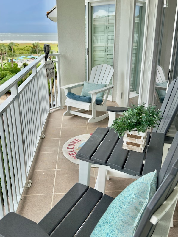 Uma varanda com vista para a praia, uma cadeira Adirondack branca, uma mesinha com uma planta e um tapete de boas-vindas.