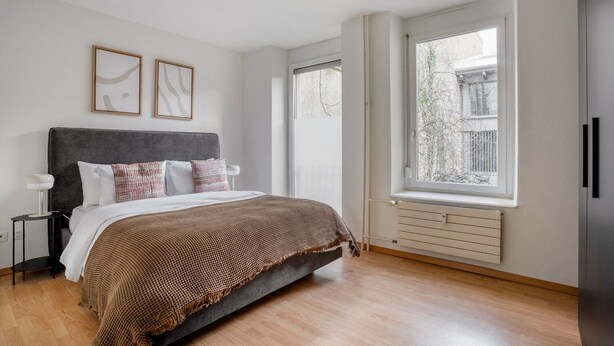 A bedroom with a bed, a bedside table, a window, and a radiator. - Stellar Matthäus 1BR w/ Laundry, 13 min to City Center, by Blueground (Basel)