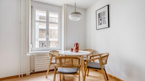 A dining area with a round table, four wicker chairs, a white ceiling light, and a window with a view of buildings. - Stellar Matthäus 1BR w/ Laundry, 13 min to City Center, by Blueground (Basel)