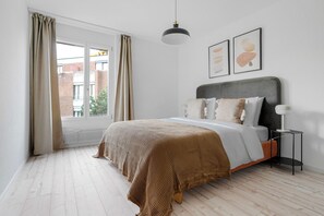 Ein Schlafzimmer mit Bett, einem Fenster mit Vorhängen, einer Pendelleuchte und zwei gerahmten Bildern an der Wand.