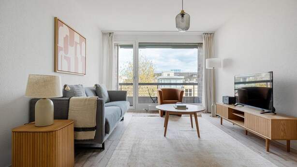 Ein modernes Wohnzimmer mit einer Couch, einem Couchtisch, einem Fernseher und einem Balkon mit Blick auf Gebäude.