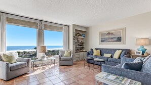 Uma sala de estar espaçosa com vista para o oceano, apresentando um sofá xadrez, um sofá azul e uma mesa de centro de vidro com revistas.