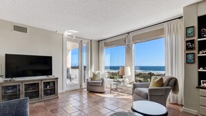 Uma sala de estar ampla, com uma TV grande, assentos confortáveis e vista para o oceano.