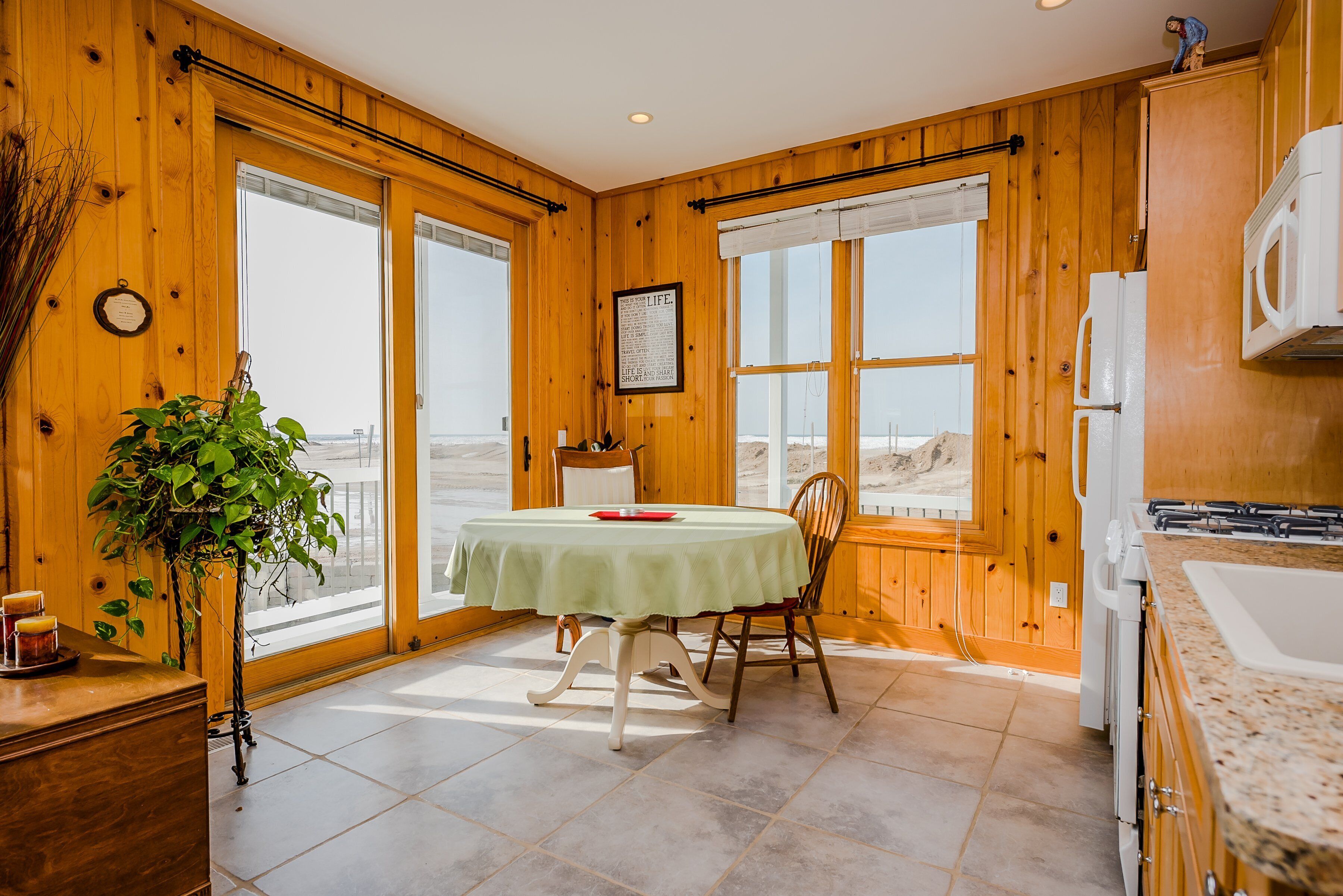 A wooden cabin interior with a dining table, chairs, a potted plant, a framed picture, and a kitchen area.