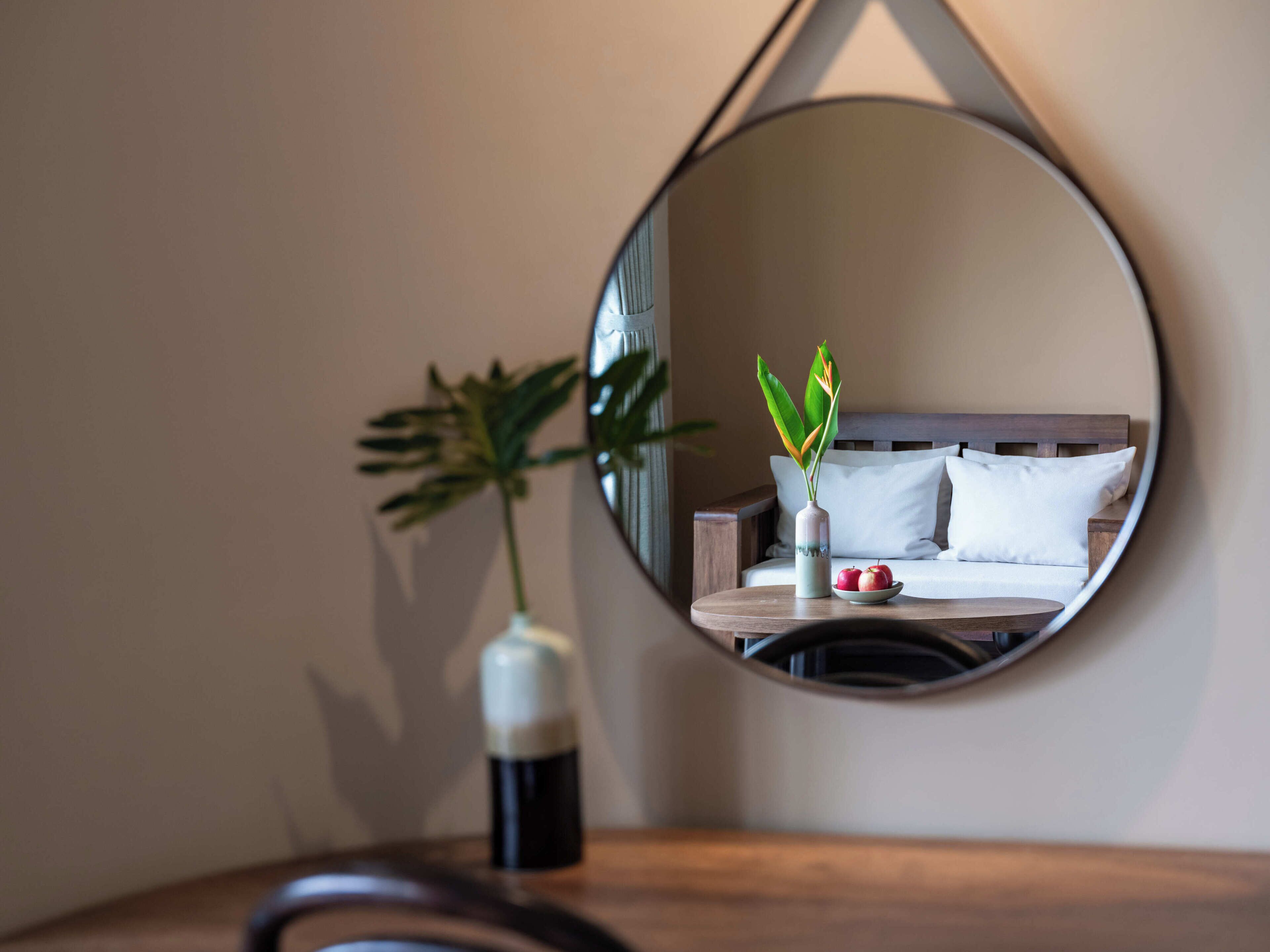 Un espejo redondo que refleja una cama, un florero con una planta y un cuenco con manzanas.