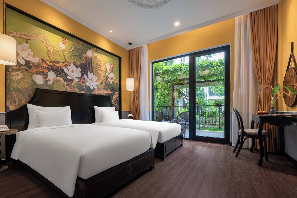 A hotel room with two beds, a balcony with a view of greenery, a dining area with a table and chairs, and a large floral painting on the wall.