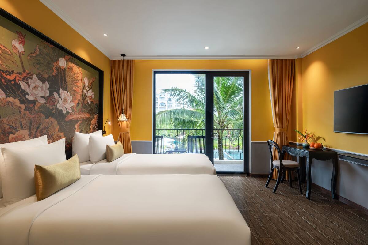 A hotel room with two beds, a desk, a TV, and a balcony with a view of palm trees.