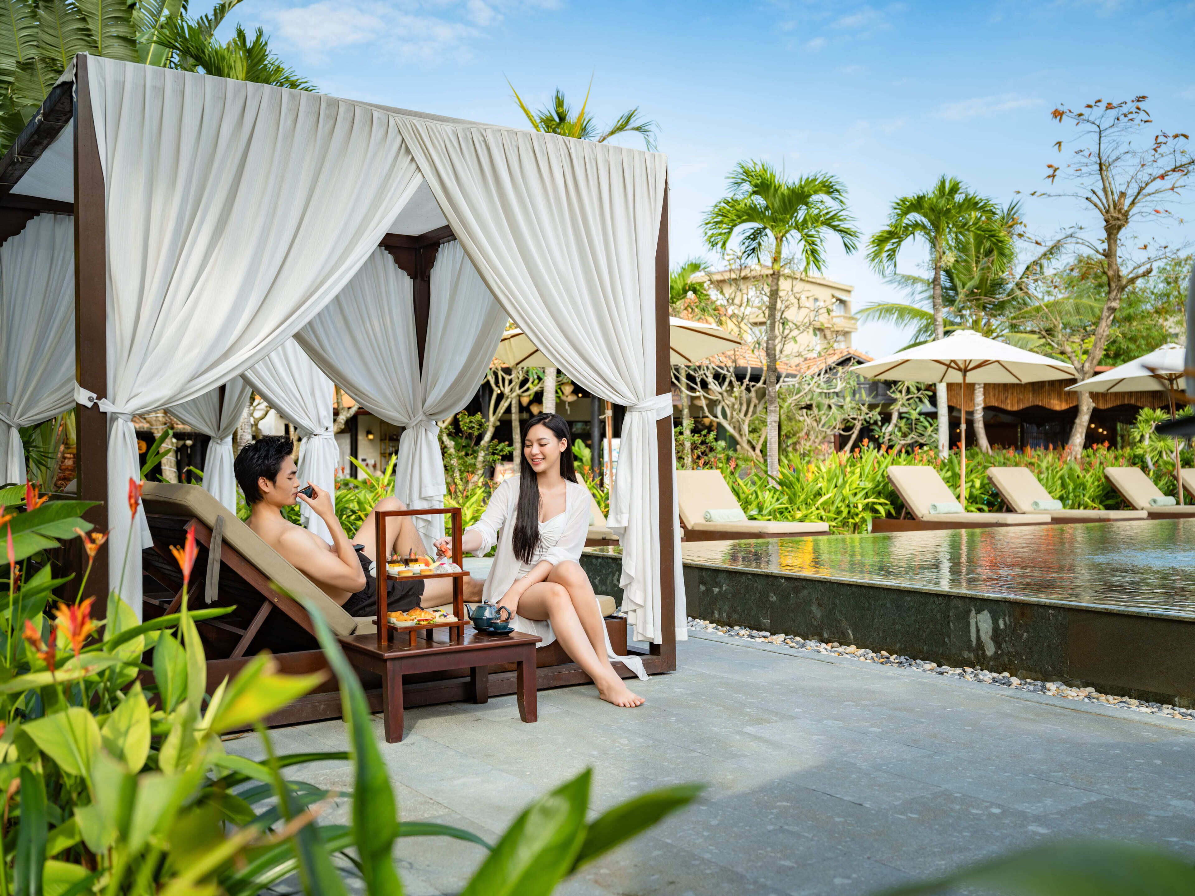 A couple relaxing under a canopy by a pool, with lounge chairs and umbrellas in the background.