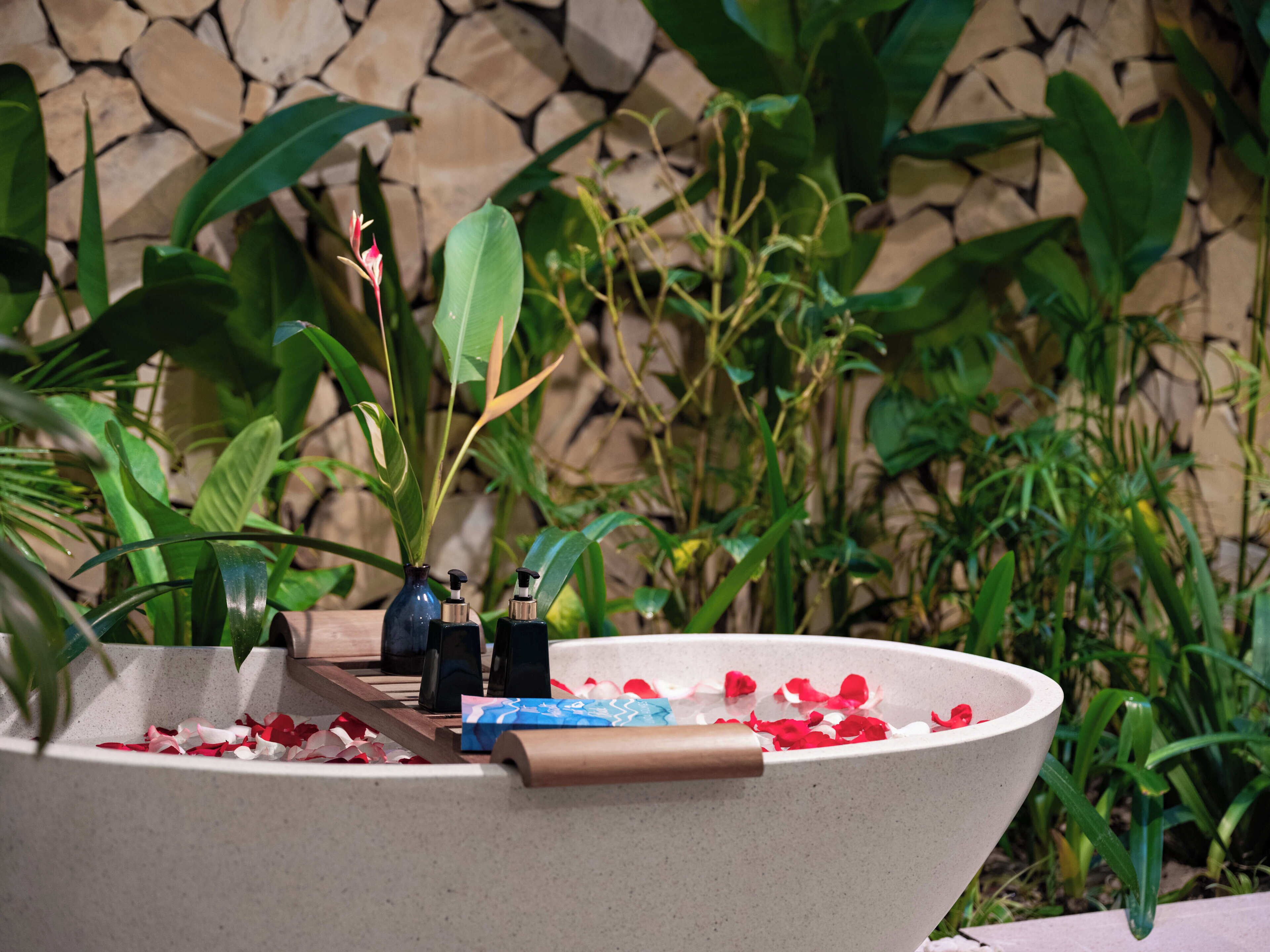 A stone wall with a large planter containing a variety of plants and a decorative bowl with rose petals and a small vase.