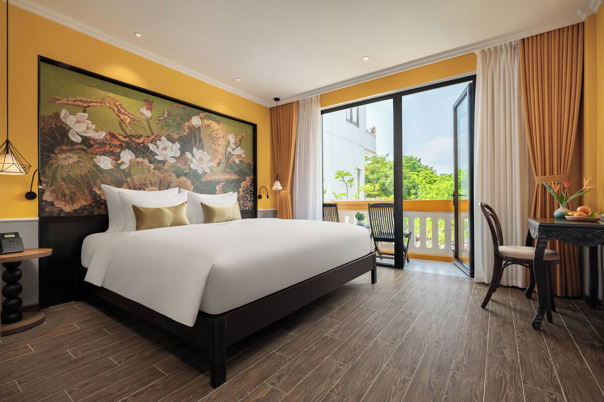 A hotel room with a large bed, a balcony with a view, a dining area, and a painting on the wall.